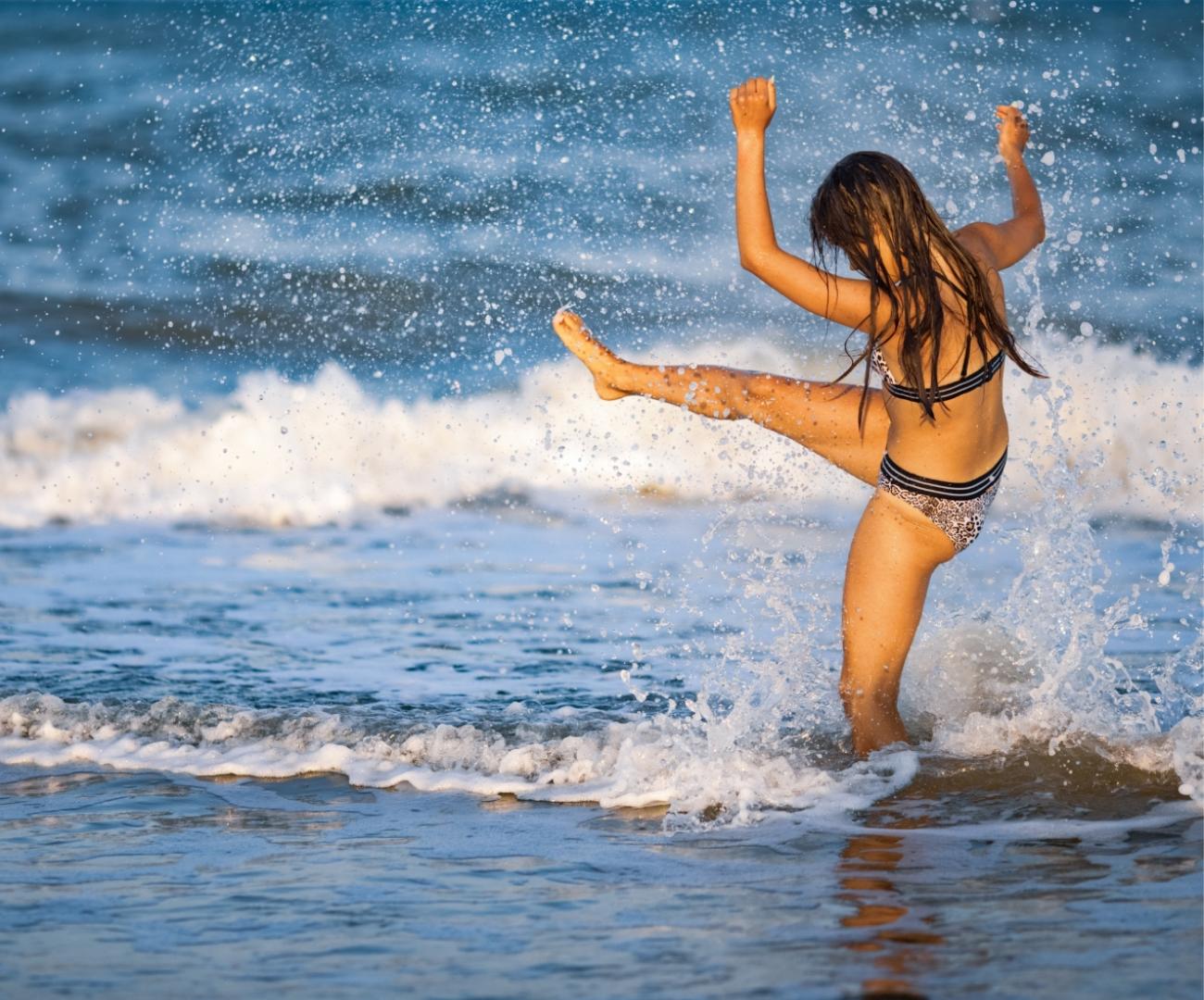 A girl plays in the water at the beach, lifting one leg.