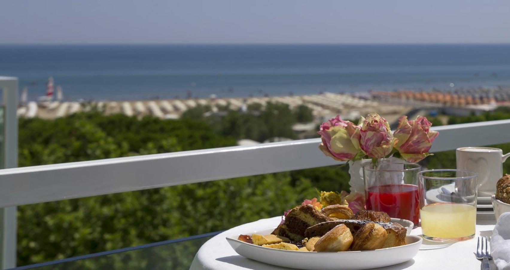 Colazione con vista mare, dolci e bevande su un balcone soleggiato.