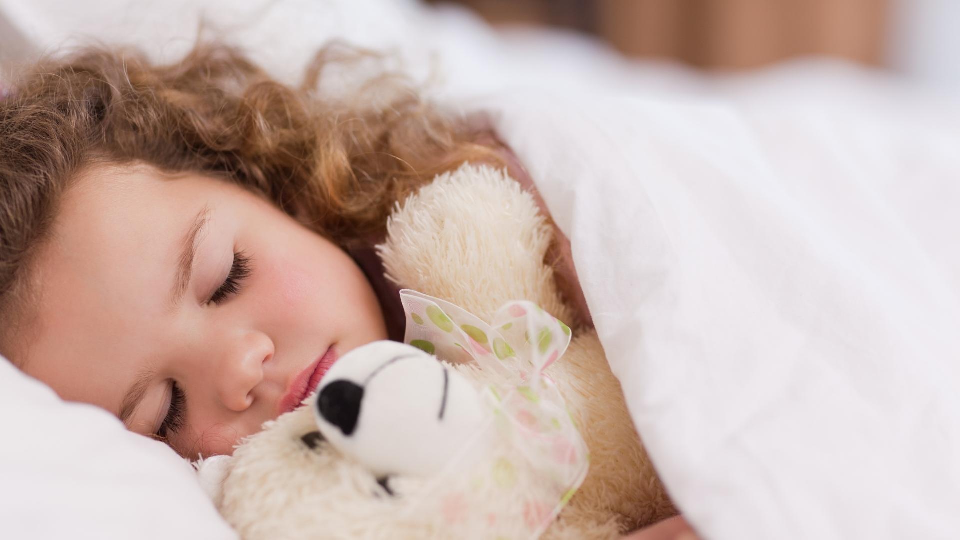 Bambina che dorme abbracciata a un peluche sotto una coperta bianca.